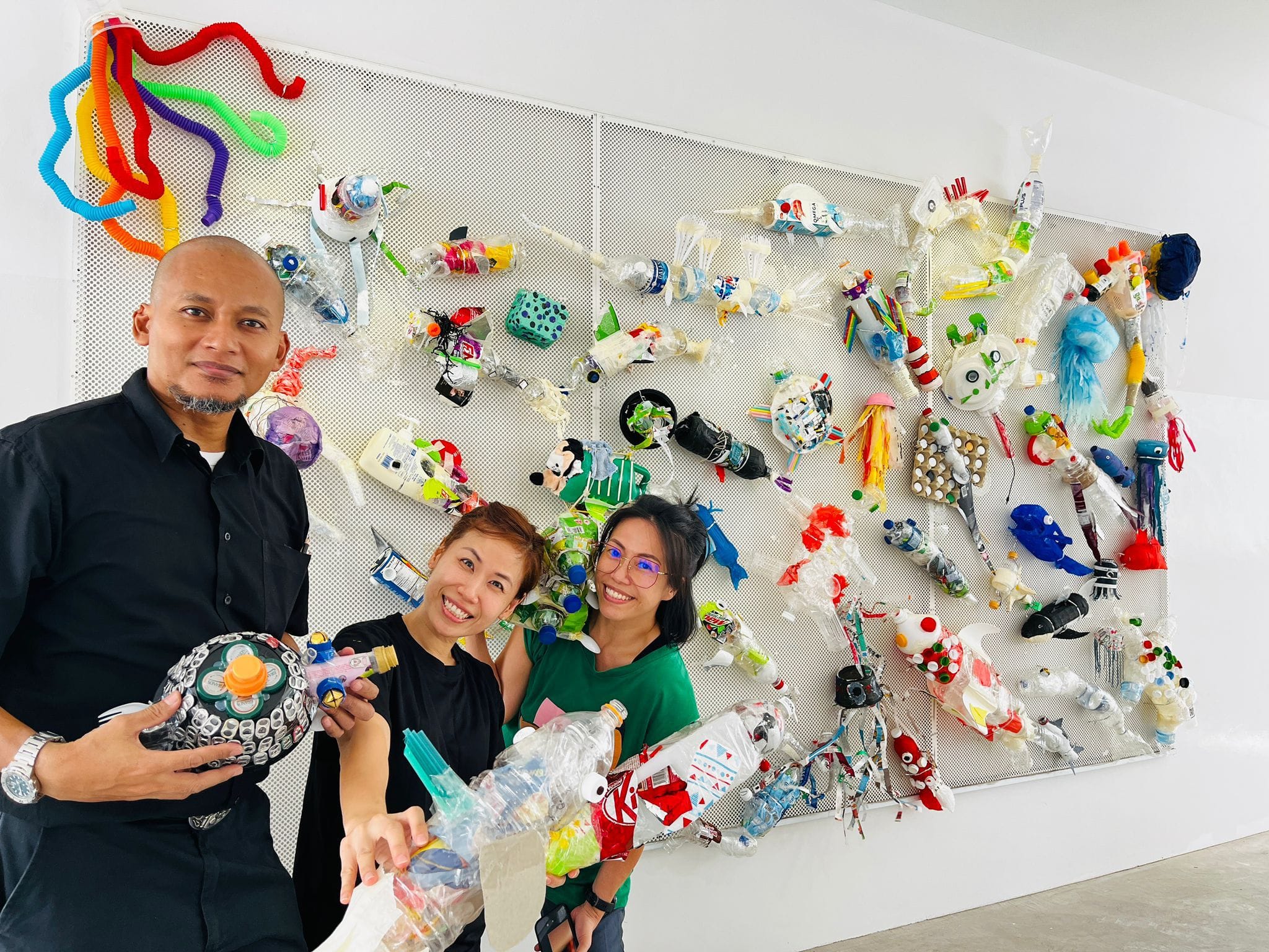 Three smiling people stand before a large white wall covered in colorful recycled plastic waste sculptures.